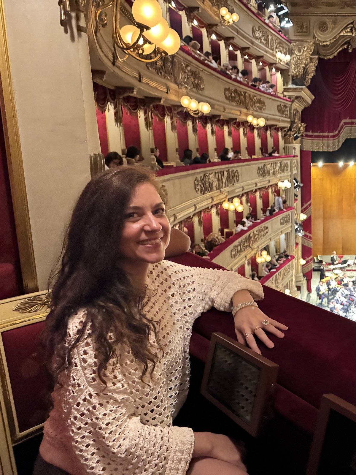 Teri smiling from a box at La Scala, in a cream knit top, with the tiers of boxes and the orchestra on stage behind her