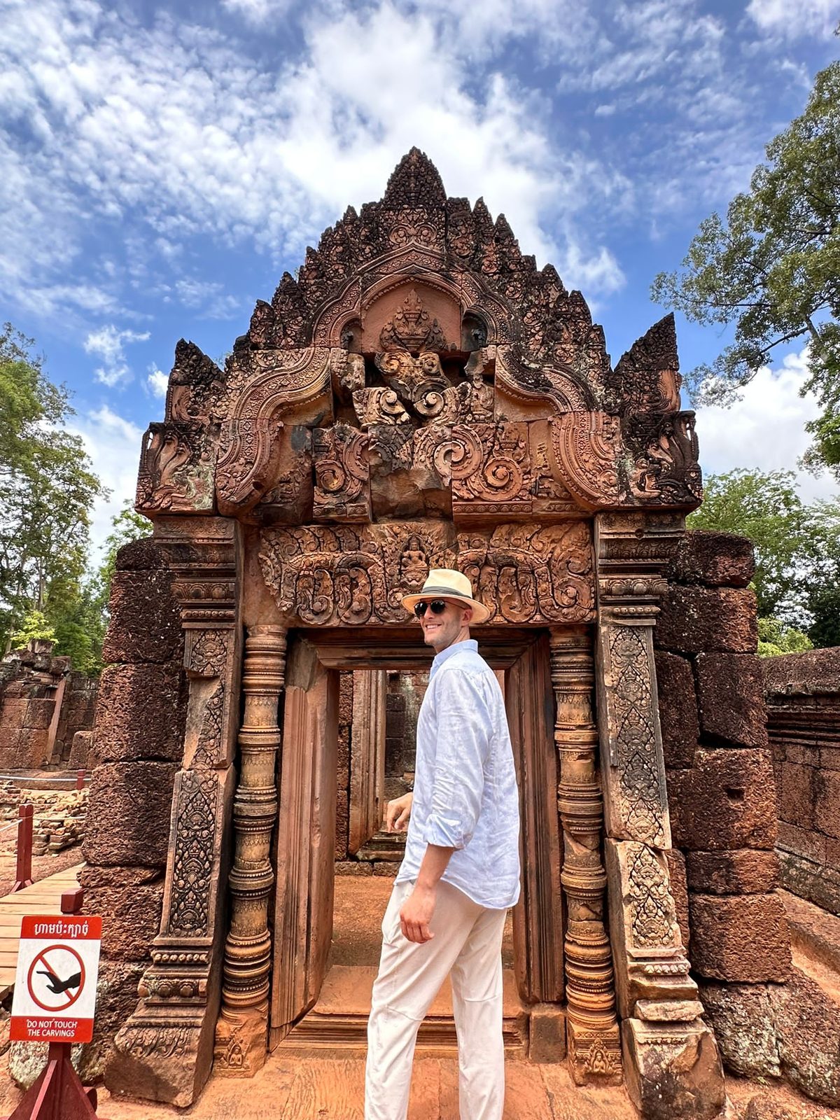 Thomas in a straw hat and a light linen shirt at a carved pink sandstone doorway at Banteay Srei