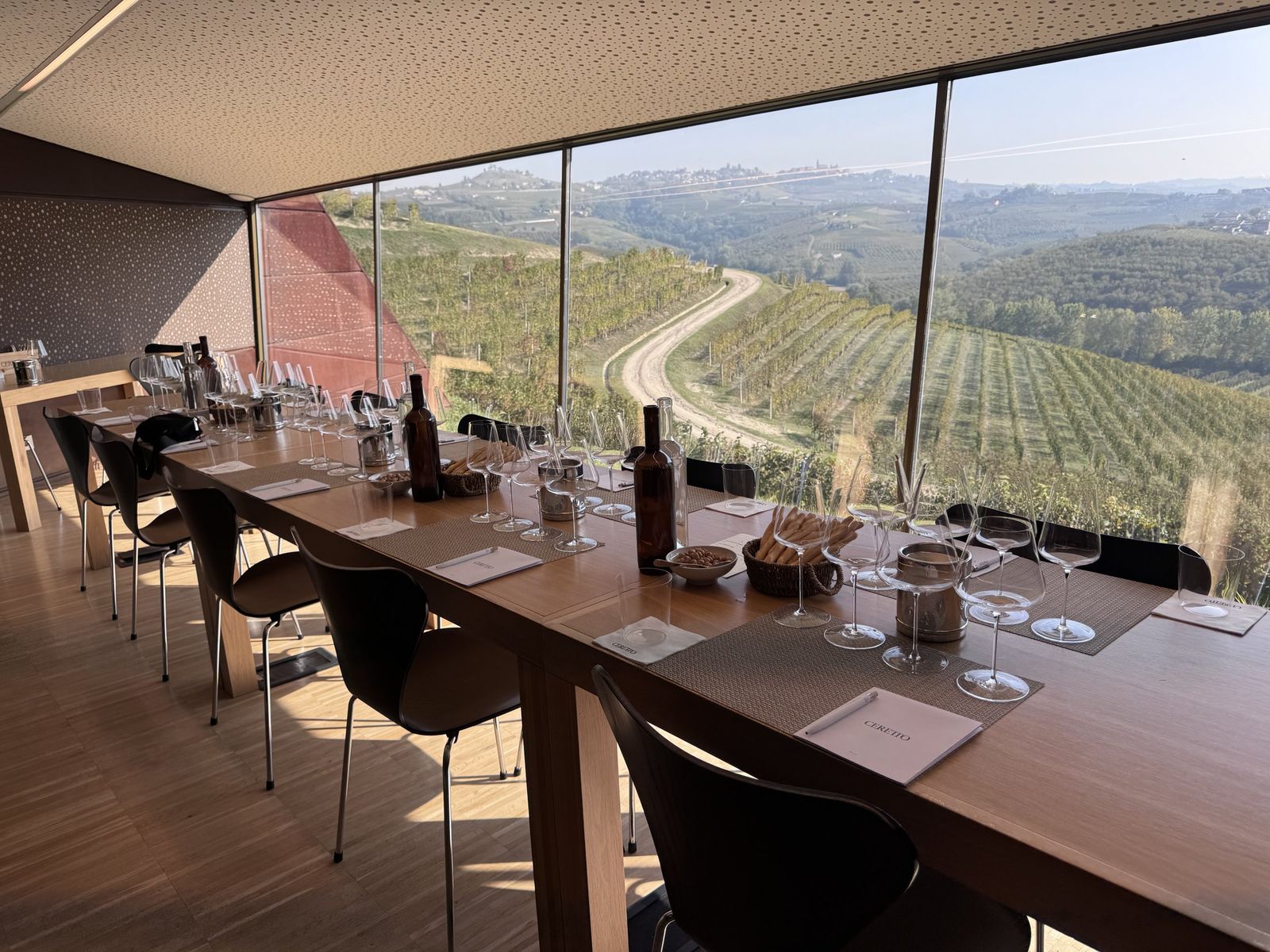 A long tasting table set with glasses at Ceretto, with a window onto vineyards and a white road winding through them