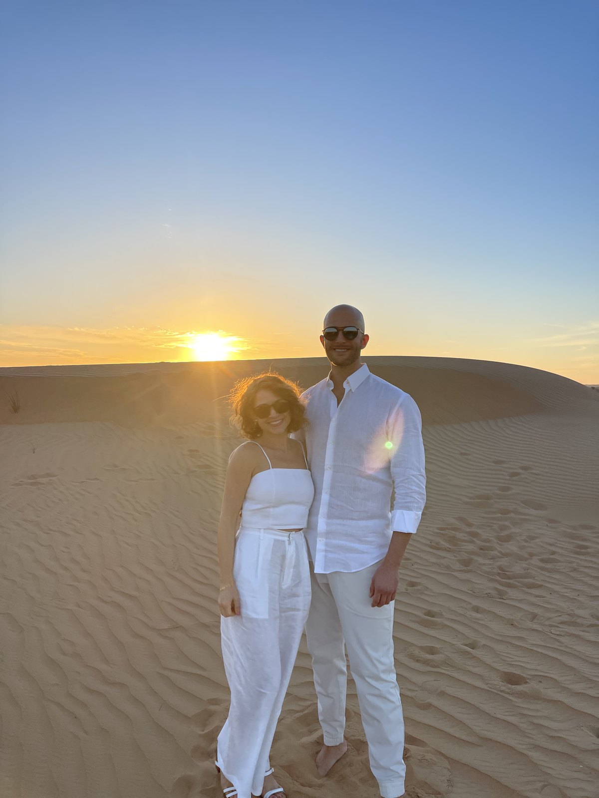 Thomas and Teri in white on a dune with the sun setting between them