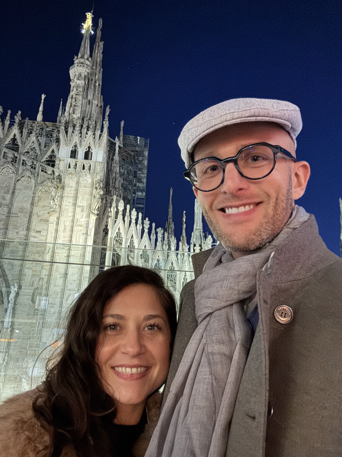 Thomas in a flat cap and Teri smiling in front of the lit spires of Milan Cathedral at night