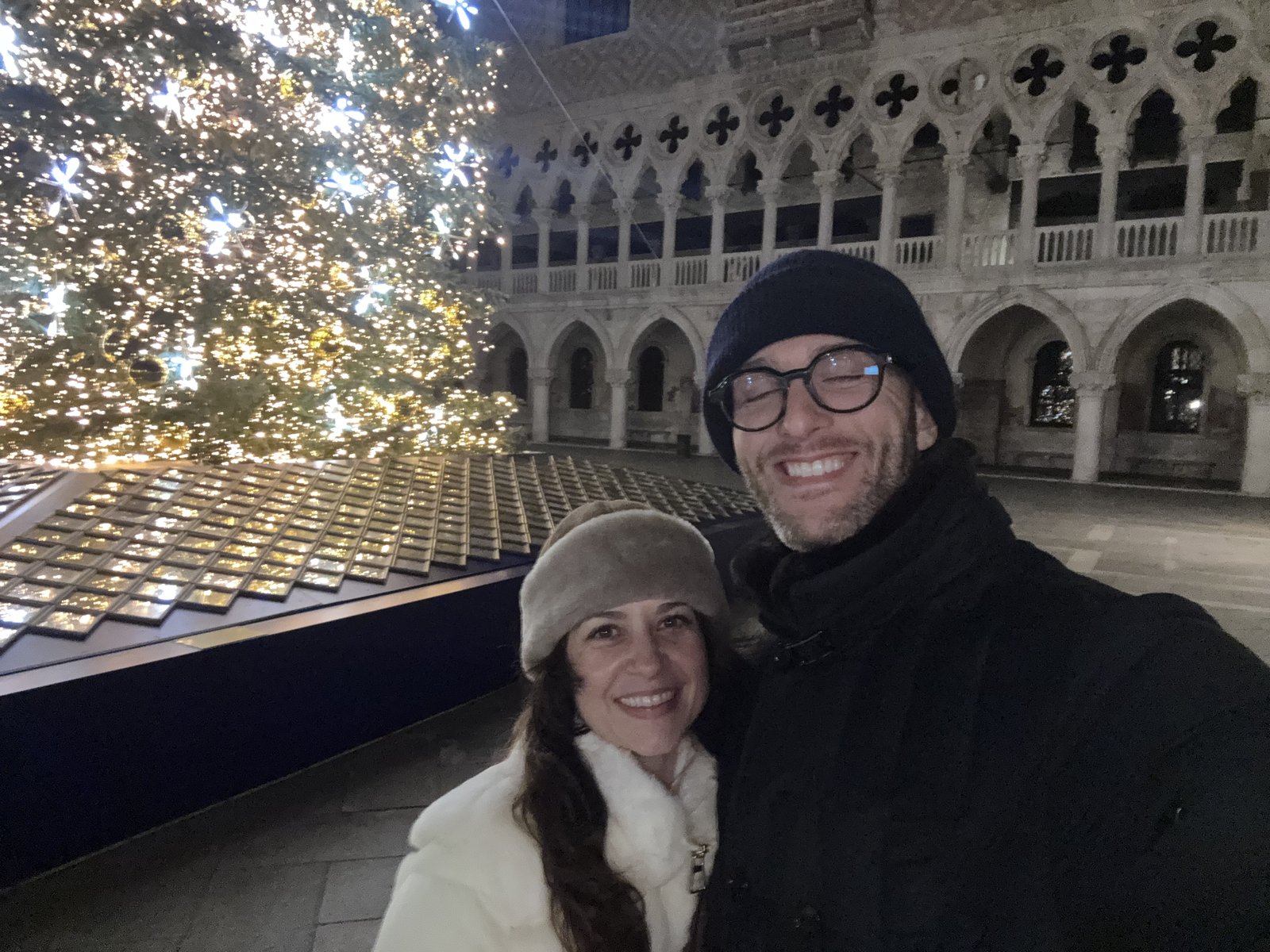 Thomas and Teri smiling in winter coats beside the lit Christmas tree, with the Doge's Palace behind them at night