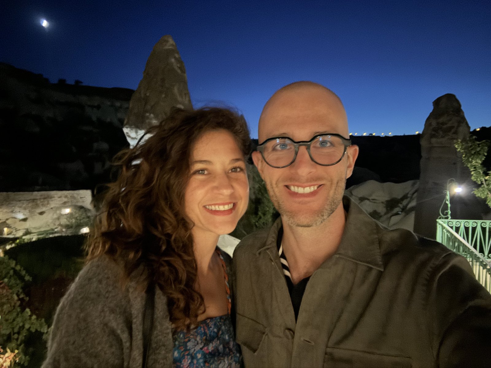 Thomas and Teri smiling at dusk, with the rock chimneys of Cappadocia and the moon behind them